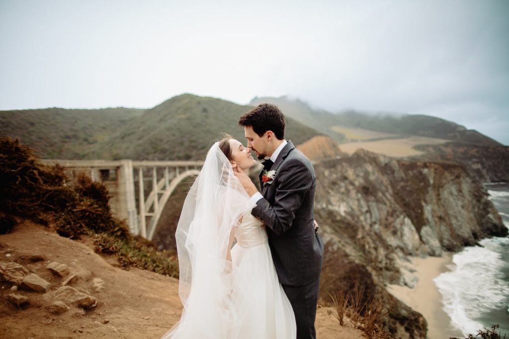A couple eloping in front of Bixby Bridge in Big Sur, California. They are facing each other, and the groom is caressing her chin and pulling her in for a kiss. 
