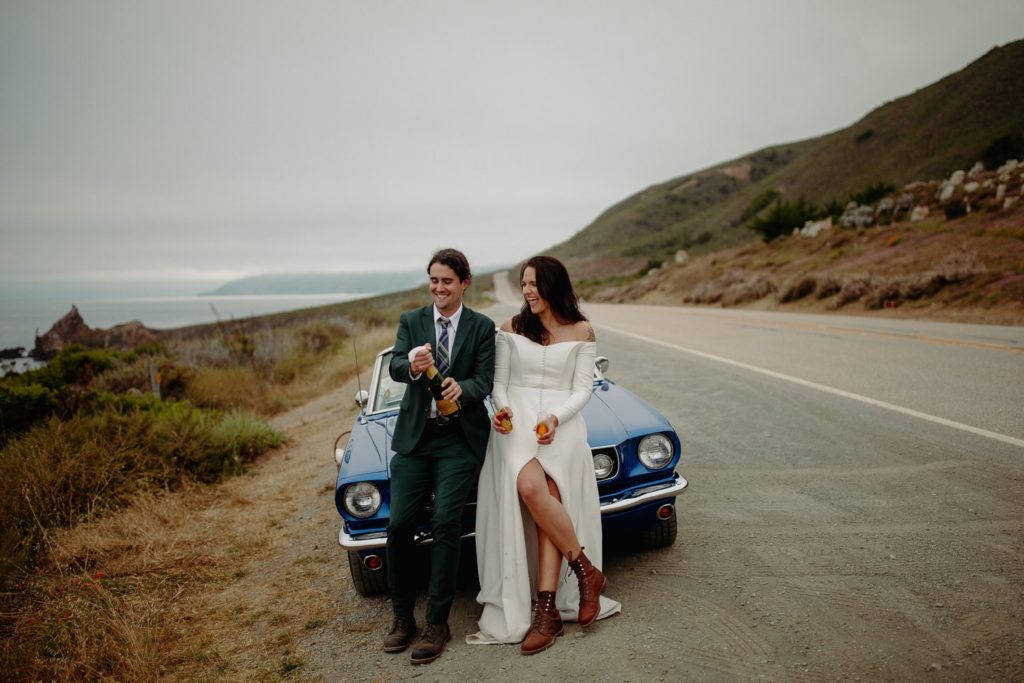 A couple popping champagne in front of a vintage car on the side of the coastal highway to celebrate their Big Sur elopement.