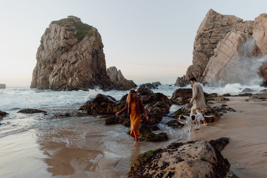A couple is walking on a beach in Portugal with their dog. The water is reaching their bare feet, and they're surrounded by rocks.