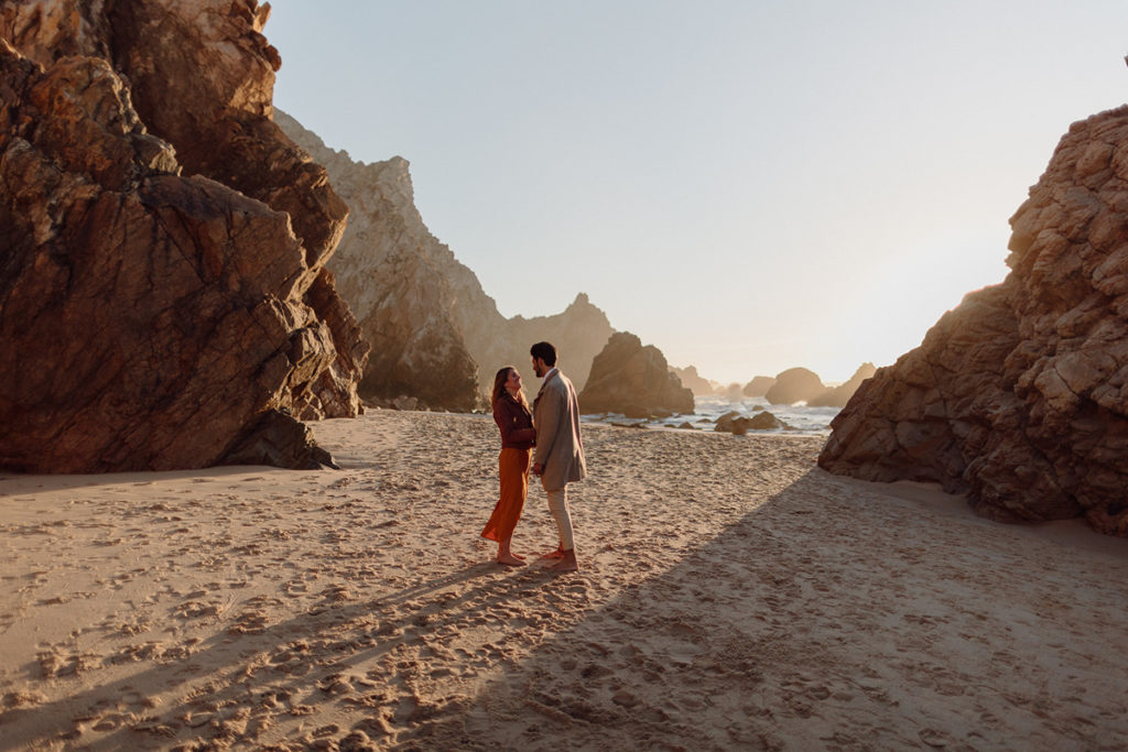 A couple is standing barefoot in the sand on a beach in Portugal, with the ocean behind them. They're surrounded by rocks, facing each other.