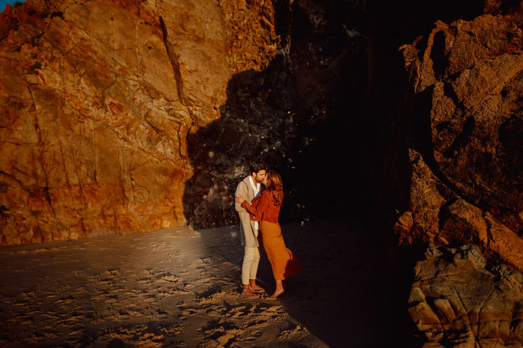 A couple is kissing on the beach in Portugal, standing barefoot in the sand. Around them is a sea cave.