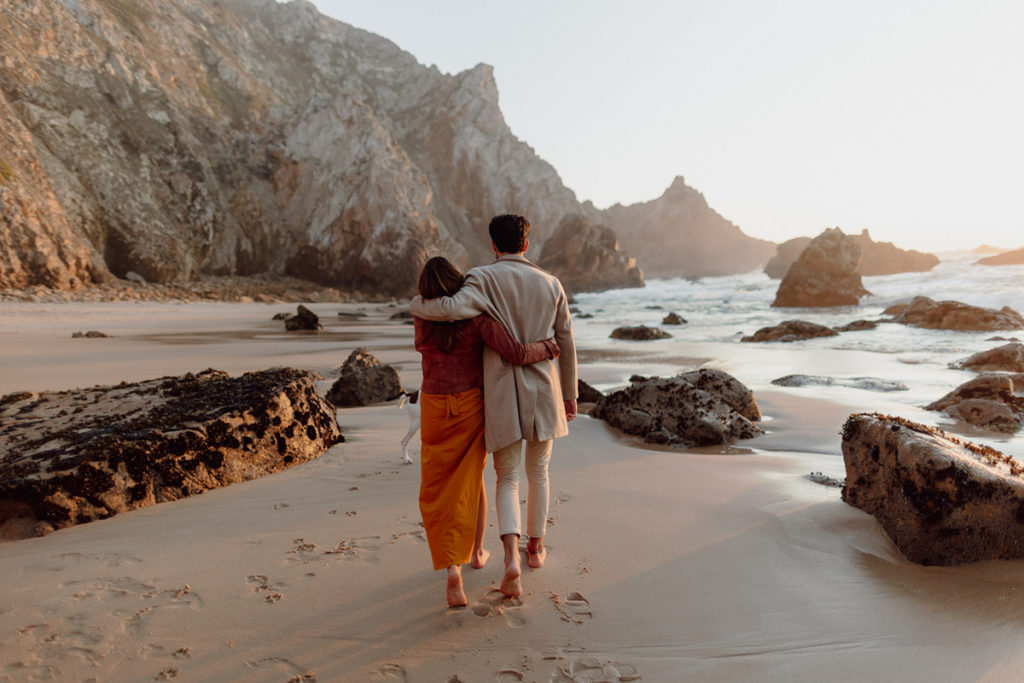 A couple is walking on a beach in Portugal, with their arms around each other. They're barefoot, surrounded by rocks. 