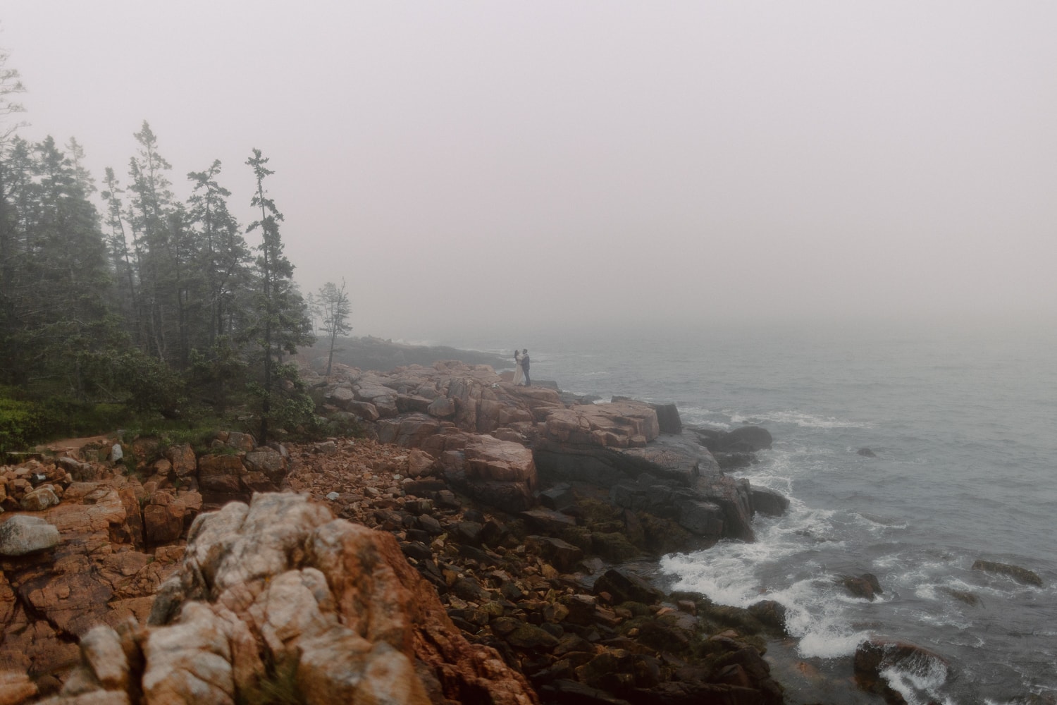 Moody elopement in Maine. Couple in wedding attire standing on rocky cliffs overlooking the ocean.