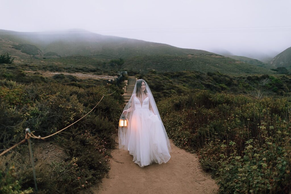 Bride with long veil and lantern walking a path in Big Sur
