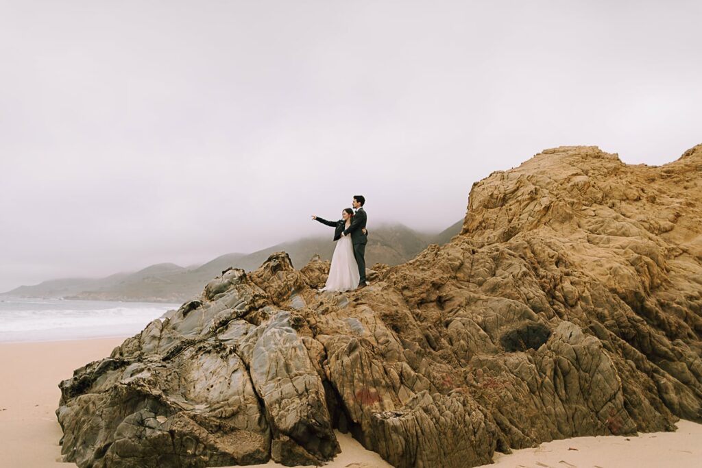 Couple standing on a large rock on the beach in Big Sur on wedding day with leather jacket