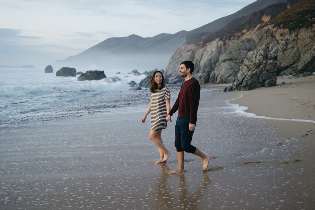 A couple is walking barefoot on the beach at Garrapata State Park, holding hands.